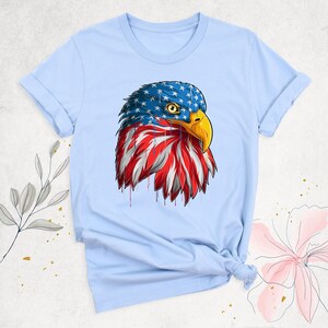 May include: Light blue t-shirt featuring a detailed illustration of an American bald eagle's head. The eagle's head is adorned with the American flag design, with stars and stripes. The eagle has a yellow beak and a focused gaze.