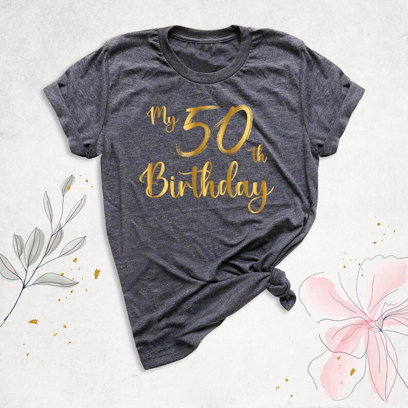 May include: A dark grey t-shirt with gold foil lettering that reads "My 50th Birthday".