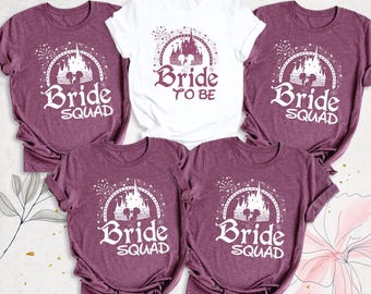 Może przedstawiać: Cztery koszulki w kolorze bordowego melanżu oraz jedna biała koszulka. Bordowe koszulki mają białą grafikę zamku z napisem Bride Squad. Biała koszulka ma bordową grafikę zamku z napisem Bride To Be.
