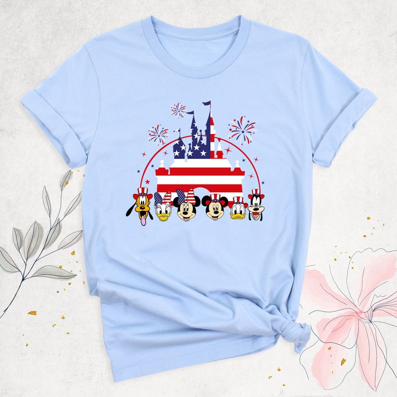 May include: Light blue t-shirt with a graphic of Mickey Mouse, Minnie Mouse, Donald Duck, Goofy, and Pluto wearing red, white, and blue hats in front of a castle with an American flag. The castle is surrounded by fireworks.