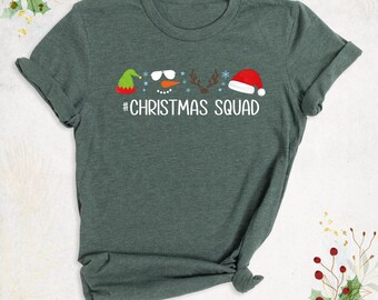 May include: A dark green short-sleeved t-shirt featuring a graphic print of a green elf hat, snowman face with sunglasses, reindeer antlers, and a red Santa hat. Below the icons, the text reads #CHRISTMAS SQUAD in white letters.