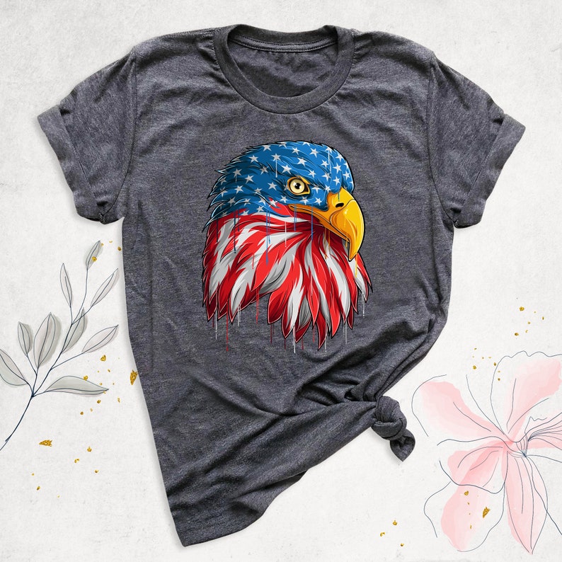 May include: Dark gray t-shirt featuring a graphic of an American bald eagle's head. The eagle's head is designed with the American flag, with blue stars on the head and red and white stripes on the chest. The eagle has a yellow beak.