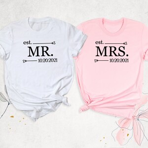 K&ouml;nnte beinhalten: Zwei wei&szlig;e T-Shirts mit schwarzem Text. Das linke Shirt sagt "est. MR. 10.20.2021" und das rechte Shirt sagt "est. MRS. 10.20.2021".