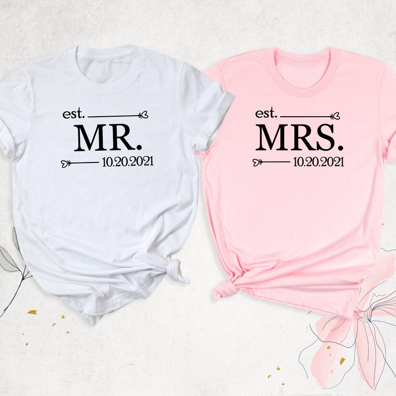 K&ouml;nnte beinhalten: Zwei wei&szlig;e T-Shirts mit schwarzem Text. Das linke Shirt sagt "est. MR. 10.20.2021" und das rechte Shirt sagt "est. MRS. 10.20.2021".