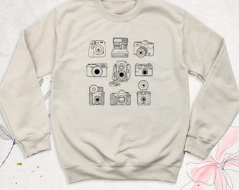 May include: A beige crewneck sweatshirt featuring a black line art graphic of nine different vintage cameras arranged in a grid. The long-sleeved garment has a ribbed collar, cuffs, and hem, showcasing various camera styles.