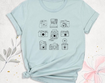 May include: A light blue short-sleeve t-shirt featuring a central graphic print of nine different vintage cameras arranged in a grid. The cameras are depicted in black line art, showcasing various styles including instant and film models.