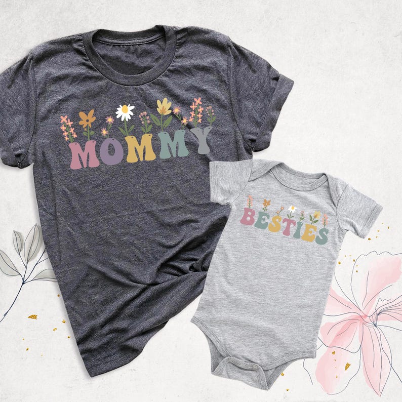 May include: Two matching t-shirts. One is a dark gray crew neck with the word "MOMMY" in colorful letters, decorated with floral accents. The other is a light gray baby onesie with the word "BESTIES" in similar colors and floral accents.