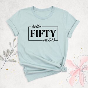 K&ouml;nnte beinhalten: Hellblaues T-Shirt mit schwarzem Text, der "hello FIFTY est. 1973" lautet.