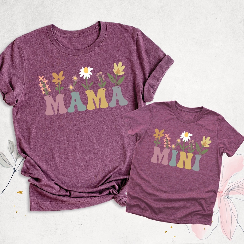 May include: Two matching maroon t-shirts. The larger shirt has the word "MAMA" in a retro font with a floral design above it. The smaller shirt has the word "MINI" with a similar floral design.