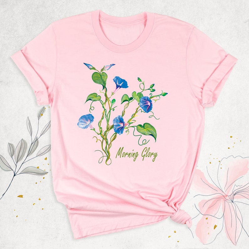 May include: A light pink t-shirt featuring a watercolor design of blue morning glory flowers and green leaves on winding vines. The words "Morning Glory" are written in green script below the flowers. The shirt has rolled-up sleeves.