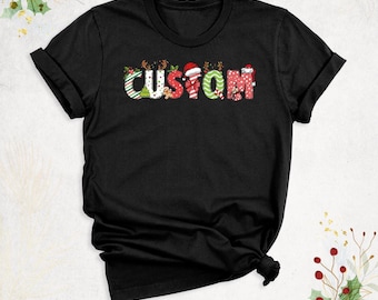 Może przedstawiać: Czarny t-shirt z krótkim rękawem z napisem CUSTOM wykonanym dużymi, stylizowanymi literami. Każda litera zawiera wzory o tematyce świątecznej, w tym laski cukrowe, poroża renifera, czapki Mikołaja, ostrokrzew i piernikowe ludziki.