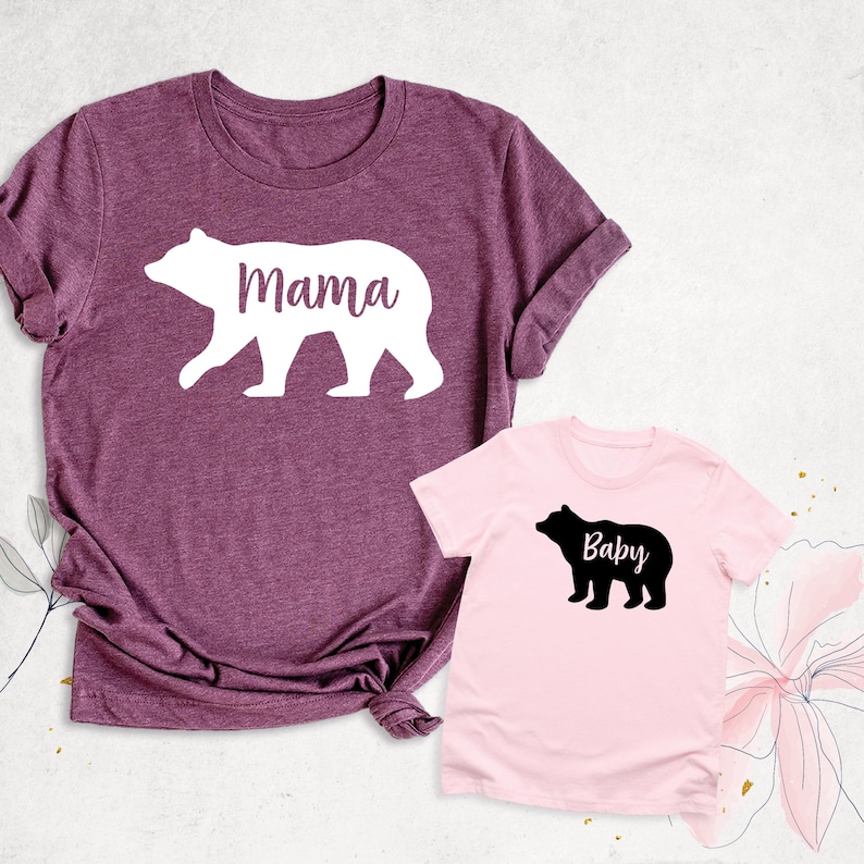 May include: A set of two t-shirts featuring a mama bear and baby bear silhouette design. The mama bear shirt is maroon with white text and a white bear silhouette. The baby bear shirt is pink with black text and a black bear silhouette. The text on the mama bear shirt says "Mama" and the text on the baby bear shirt says "Baby".