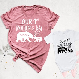 Puede incluir: Dos camisetas con un dise&ntilde;o de familia de osos. La camiseta m&aacute;s grande es rosa y dice "Nuestro 1er D&iacute;a de la Madre 2022" con una osa y un osito. La camiseta m&aacute;s peque&ntilde;a es blanca y dice "Nuestro 1er D&iacute;a de la Madre 2022" con una osa y un osito.