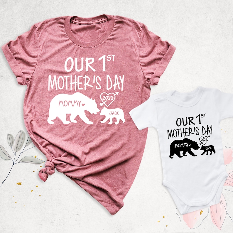 Puede incluir: Dos camisetas con un dise&ntilde;o de familia de osos. La camiseta m&aacute;s grande es rosa y dice "Nuestro 1er D&iacute;a de la Madre 2022" con una osa y un osito. La camiseta m&aacute;s peque&ntilde;a es blanca y dice "Nuestro 1er D&iacute;a de la Madre 2022" con una osa y un osito.