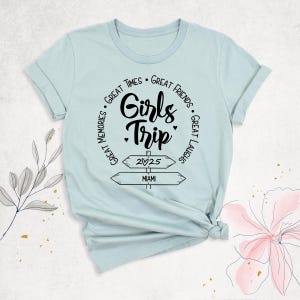 May include: A light blue t-shirt with rolled-up sleeves. The shirt features a black graphic that says "Girls Trip" with the words "Great Times, Great Friends, Great Laughs, Great Memories" around the main text. Below the text are two directional signs pointing to "2025" and "Miami".