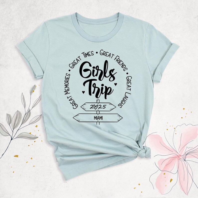 May include: A light blue t-shirt with rolled-up sleeves. The shirt features a black graphic that says "Girls Trip" with the words "Great Times, Great Friends, Great Laughs, Great Memories" around the main text. Below the text are two directional signs pointing to "2025" and "Miami".