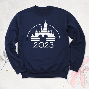 May include: Navy blue sweatshirt with a white graphic of the Disney castle silhouette, Mickey Mouse ears, and the year 2023.