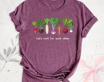 May include: A heathered maroon short-sleeve t-shirt featuring a graphic of various colorful root vegetables including radishes, a carrot, and a turnip. Below the vegetables, the text reads, Let's root for each other in white letters.