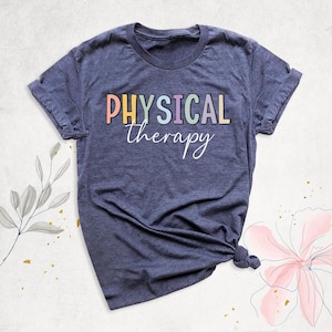 May include: A heather navy blue short-sleeve t-shirt featuring the word PHYSICAL in colorful block letters and the word therapy in white cursive script centered on the chest. The shirt has a crew neckline and a casual fit.