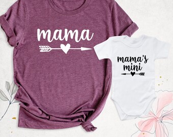 May include: A heathered maroon t-shirt with the white text "mama" above a white arrow and heart graphic, paired with a white baby bodysuit featuring the black text "mama's mini" above a black arrow and heart graphic.