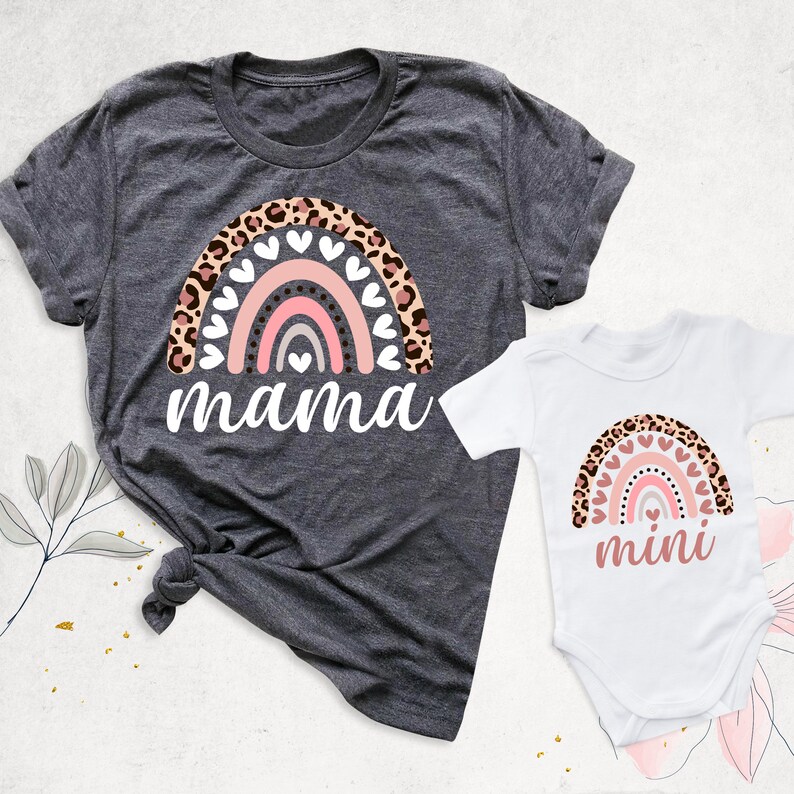 Puede incluir: Un conjunto a juego de camisetas con un arco&iacute;ris con rayas rosas, grises y blancas, rodeado de estampado de leopardo y corazones blancos. La camiseta m&aacute;s grande dice "mama" y la m&aacute;s peque&ntilde;a dice "mini".