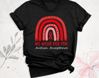 May include: A black short-sleeved t-shirt featuring a graphic of a rainbow composed of red and pink arches. Centered within the rainbow is a red awareness ribbon. Below the graphic, the text reads WE WEAR RED FOR Autism Acceptance.