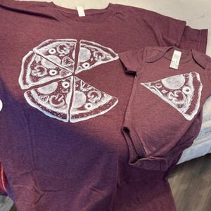 以下が含まれることがあります： マルーン色の霜降り半袖Tシャツと、お揃いのベビー用ボディスーツ。Tシャツには1ピース欠けたピザの白いグラフィックが描かれており、ボディスーツにはその欠けた1ピースのピザが白でプリントされています。