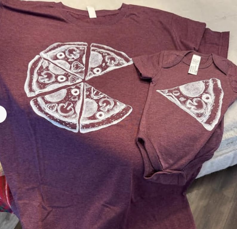 以下が含まれることがあります： マルーン色の霜降り半袖Tシャツと、お揃いのベビー用ボディスーツ。Tシャツには1ピース欠けたピザの白いグラフィックが描かれており、ボディスーツにはその欠けた1ピースのピザが白でプリントされています。