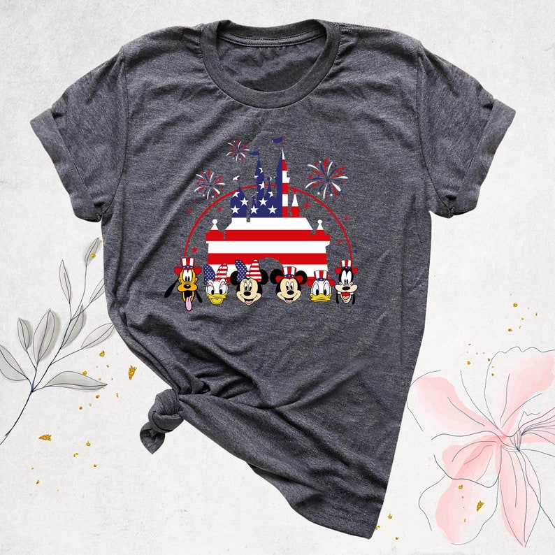 May include: A gray t-shirt with a graphic of Mickey Mouse, Minnie Mouse, Donald Duck, Goofy, and Pluto in front of a castle with an American flag. The characters are wearing red, white, and blue hats. There are fireworks in the background.