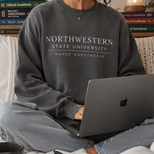 Könnte beinhalten: Ein dunkelgraues Sweatshirt mit dem weißen Text "NORTHWESTERN STATE UNIVERSITY NURSE ANESTHESIA". Die Person trägt das Sweatshirt und benutzt einen Laptop. Bücher und ein Notizbuch sind ebenfalls sichtbar.