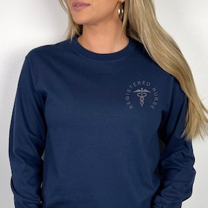 Op de afbeelding: Een marineblauw longsleeve t-shirt met de tekst "Registered Nurse" en een caduceus symbool in een cirkel op de rug.