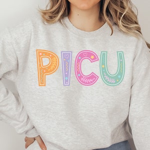 Puede incluir: Una sudadera gris claro con la palabra "PICU" impresa en la parte delantera en letras de colores con detalles florales y estrellas.