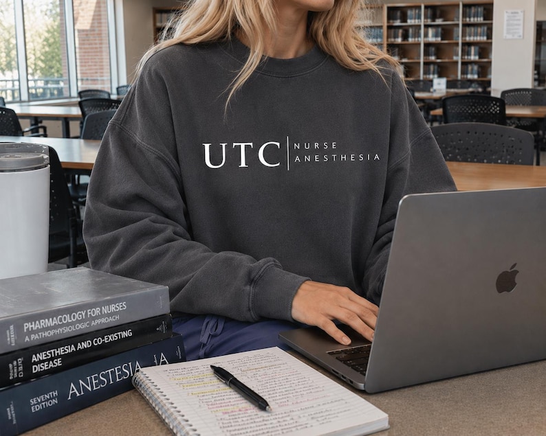 May include: Dark grey sweatshirt with the text "UTC NURSE ANESTHESIA" in white. A laptop, books, and a notebook with a pen are on the desk. The books are titled "Pharmacology for Nurses", "Anesthesia and Co-existing Disease", and "Anestesia".