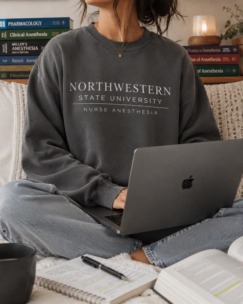 May include: A dark gray sweatshirt with the text "NORTHWESTERN STATE UNIVERSITY NURSE ANESTHESIA" in white. The person is wearing the sweatshirt and using a laptop. Books and a notebook are also visible.