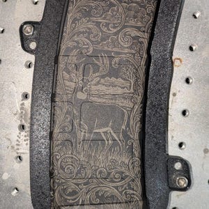 May include: A black magazine with an engraved design featuring a deer in a field, surrounded by ornate floral patterns. The magazine is set within a black frame, and the text "5.56 X 45" is visible.