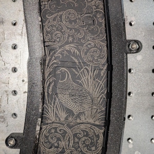 May include: A black magazine with an engraved design featuring a quail in tall grass, surrounded by ornate scrollwork. The magazine is set within a black frame, mounted on a metal surface.