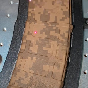 Puede incluir: Un cargador de arma de fuego de color camuflaje marrón y beige. El cargador está en un soporte de plástico negro y gris.