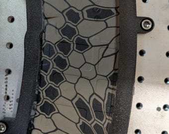 May include: A black and tan magazine with a geometric pattern is set within a black frame. The magazine has a textured surface and is secured to a metal surface with screws. The pattern is a series of interconnected shapes.
