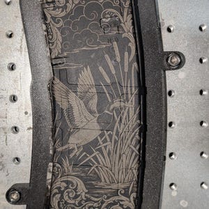 May include: A black magazine with an engraved design featuring a duck in flight over a marsh scene. The magazine is set within a black frame, mounted on a metal surface with screw holes.