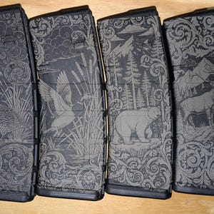 May include: Four black magazines with engraved wildlife scenes. Each magazine features a different design: a quail, a duck, a bear in a forest, and a deer with mountains. The designs are detailed and surrounded by decorative scrollwork.