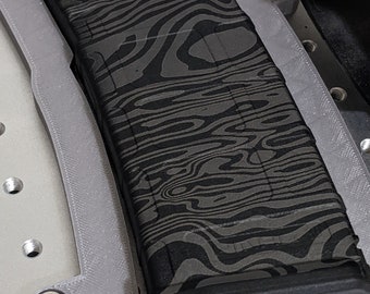 May include: A black and gray patterned magazine sits in a gray plastic mold. The mold has a curved shape and is designed to hold the magazine securely.