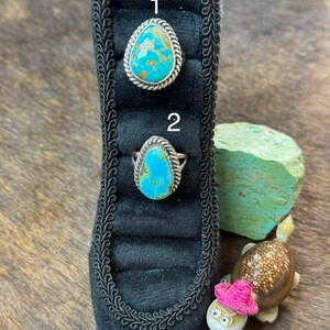 May include: Two silver rings featuring teardrop-shaped turquoise stones with brown matrix, each set in a twisted rope border. The rings are labeled 1 and 2 and are displayed on a black velvet ring holder shaped like a high-heeled shoe.