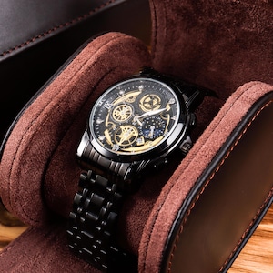 Könnte beinhalten: Eine schwarze Armbanduhr mit detailliertem gold-schwarzem Zifferblatt, präsentiert in einem braunen Uhrenetui. Die Uhr hat ein schwarzes Metallarmband und wird auf einer Holzoberfläche präsentiert. Das Zifferblatt der Uhr weist mehrere Zifferblätter und komplizierte Designs auf.