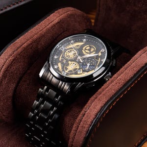 Könnte beinhalten: Eine schwarze Armbanduhr mit einem detaillierten Zifferblatt mit goldenen Akzenten und mehreren Hilfszifferblättern. Die Uhr hat ein schwarzes Metallarmband und wird in einem braunen, mit Wildleder gefütterten Uhrenetui präsentiert. Das Etui ist aus braunem Leder.