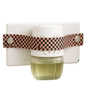 Può includere: Un diffusore d'aria bianco con una bottiglia di vetro trasparente contenente un liquido verde chiaro. Una cinghia decorativa con un motivo a scacchi neri e rosa fissa l'unità. Il diffusore d'aria è progettato per essere collegato a una presa a muro.