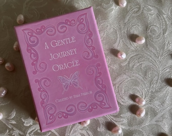 May include: A pink box with a white ornate design and the text "A Gentle Journey Oracle" and "Created by Sari Parris". A butterfly is in the center of the box.