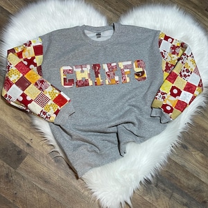 May include: A gray sweatshirt with the word "CHIEFS" spelled out in red, yellow, and white patchwork fabric. The sleeves are made of the same patchwork fabric.