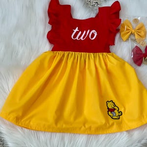May include: A red and yellow dress with the word "two" in white script. The dress has a yellow skirt and a red bodice with ruffled sleeves. A cartoon bear is embroidered on the skirt. Two hair bows are visible.
