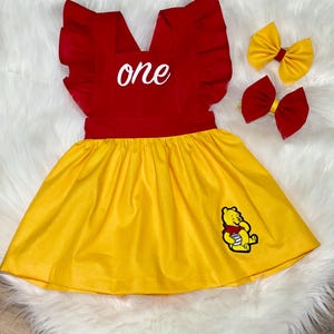 May include: A yellow and red dress with a Winnie the Pooh character on the skirt. The dress has a ruffled neckline and says "one" in white lettering. Two matching hair bows are included, one red and one yellow.