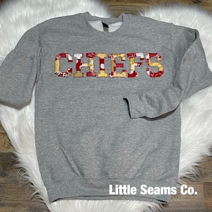 May include: Gray sweatshirt with the word "CHIEFS" in red, yellow, and white patchwork letters. The letters are outlined in yellow. The sweatshirt has a crew neck and long sleeves. The text "Little Seams Co." is printed on the bottom hem.